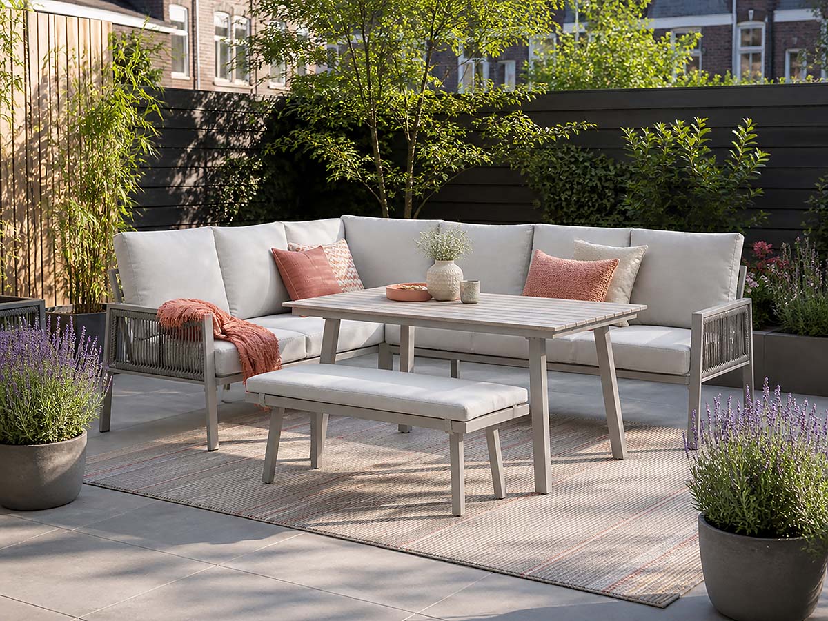 Outdoor patio furniture set with a sectional sofa, matching bench, rectangular dining table, and a woven rug in a landscaped backyard setting.